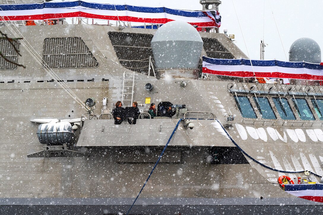 A group of people stand on the balcony of a military vessel as snow falls under a cloudy sky.