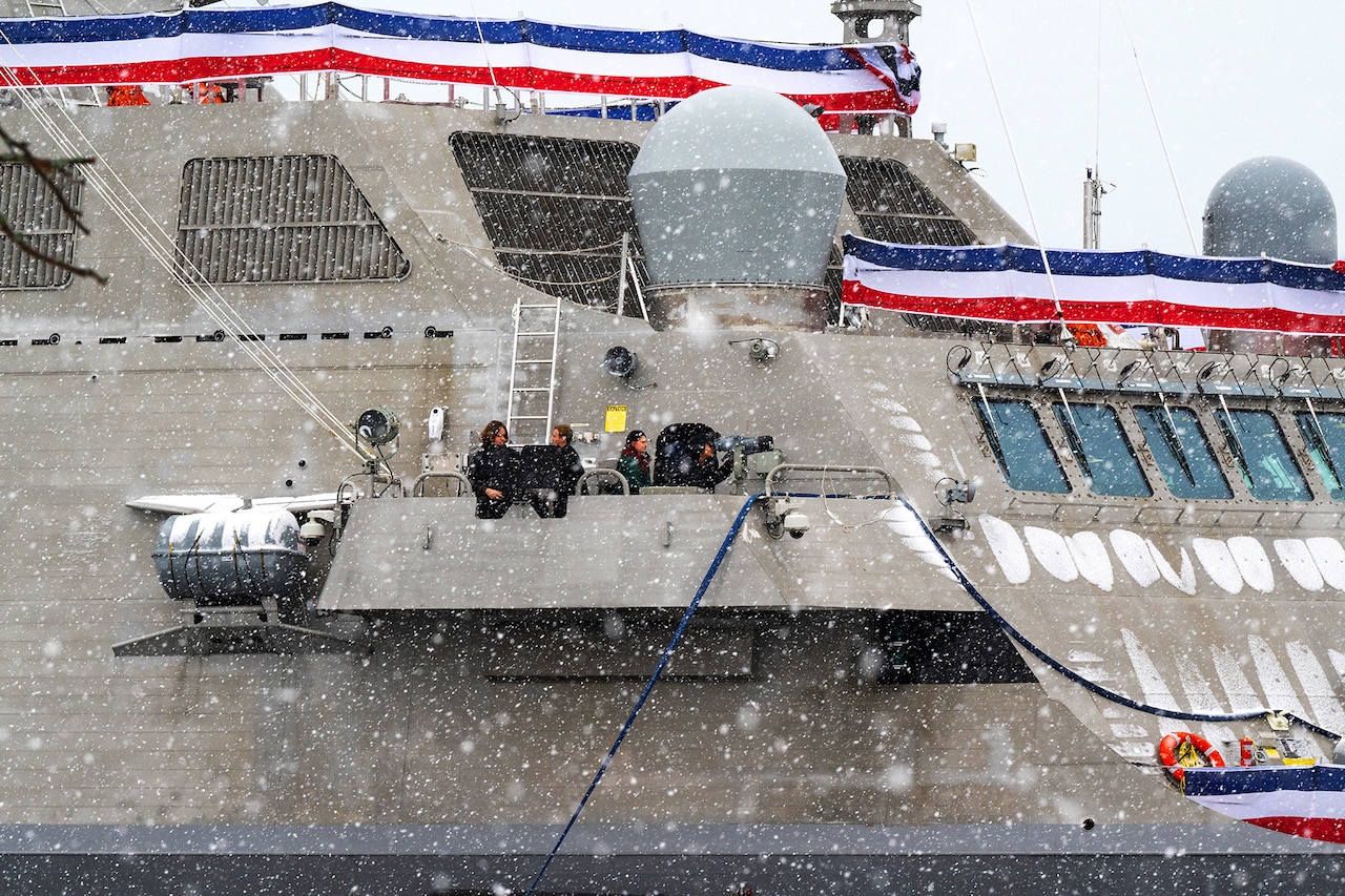 A group of people stand on the balcony of a military vessel as snow falls under a cloudy sky.