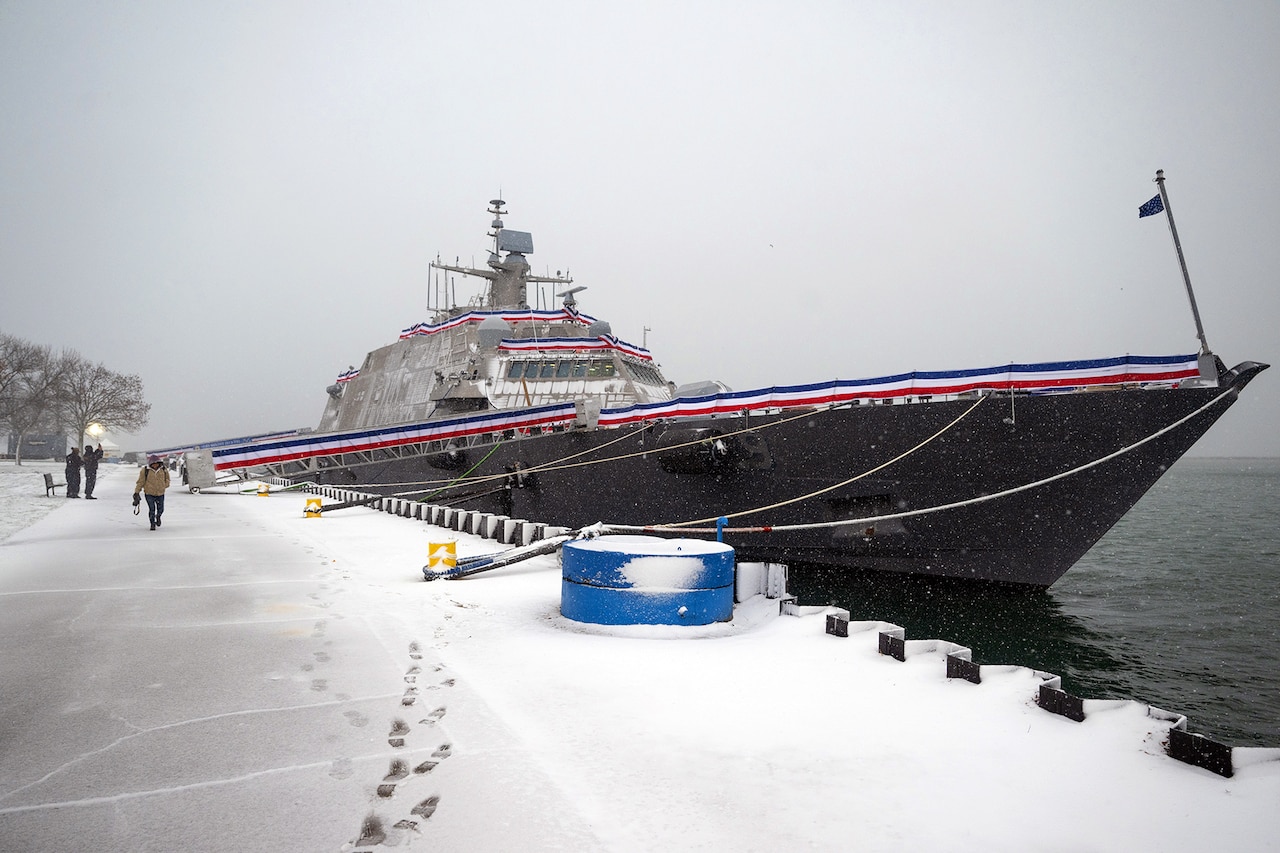A Navy vessel is parked by a snowy dock under a cloudy sky.