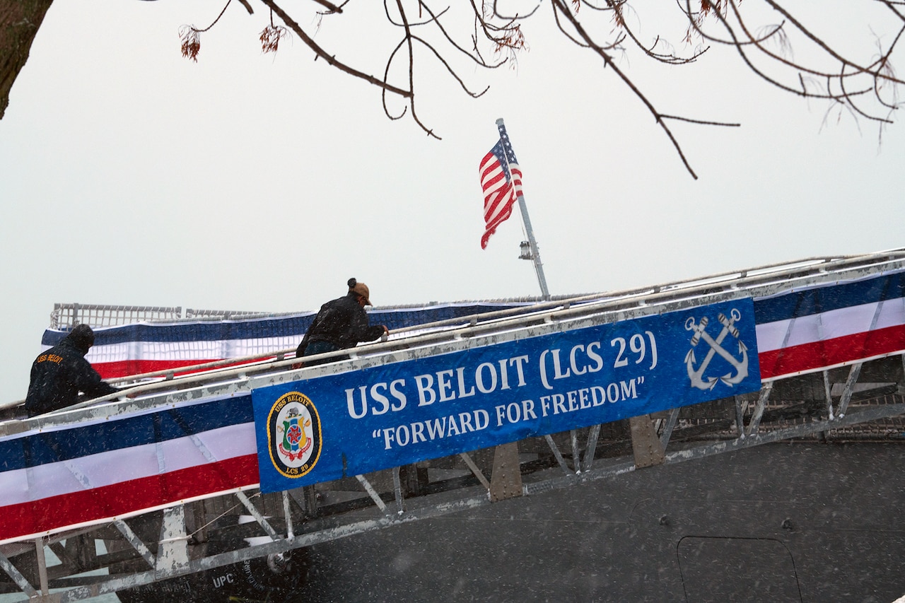 Two service members use a ramp to board a Navy vessel during a cloudy day. Banner text reads: USS Beloit, forward for freedom.