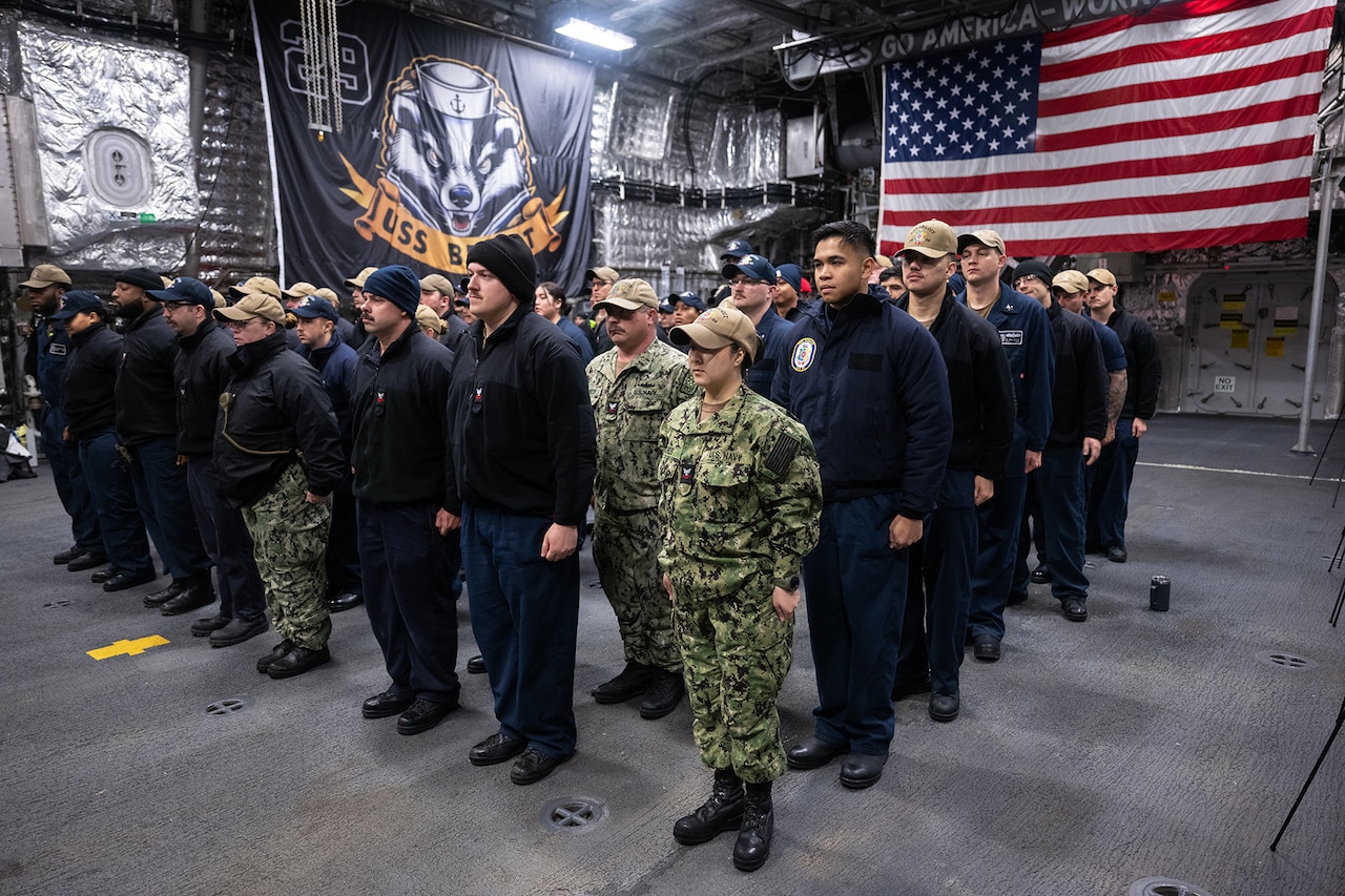 A group of sailors stand in formation inside a room with the flags of the U.S. and USS Beloit.