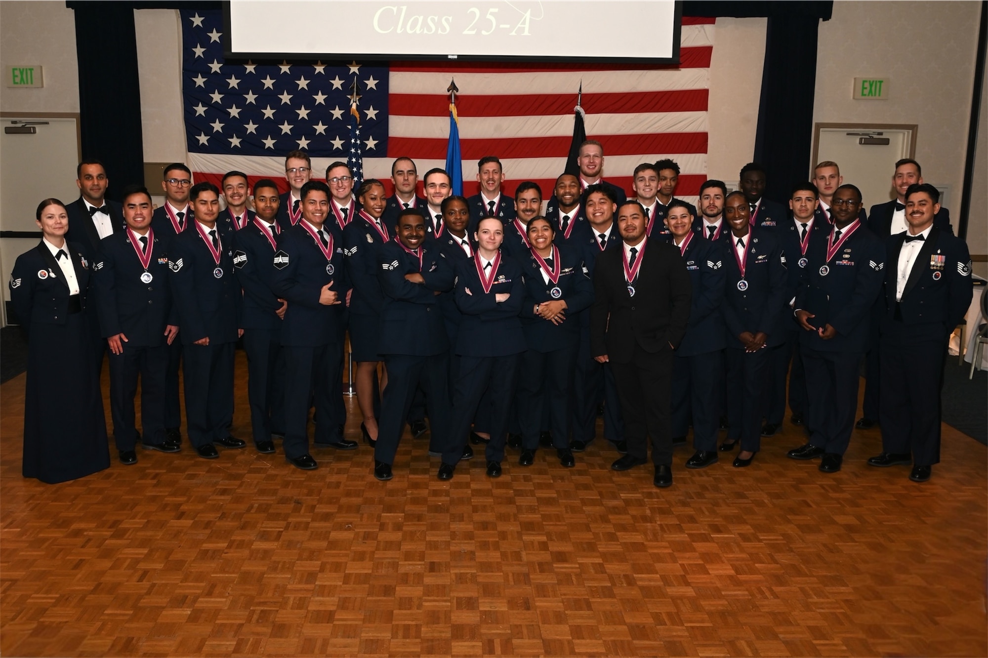 vandenberg members posing for a group photo in their blues after graduating airman leadership school, wooden floor, american flag in background