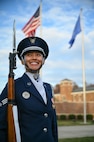 Photo of a U.S. Air Force honor guardsman smiling while holding her ceremonial rifle outside. The U.S. and Air Force flags wave in the background.