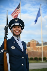 Photo of a U.S. Air Force honor guardsman smiling while holding her ceremonial rifle outside. The U.S. and Air Force flags wave in the background.