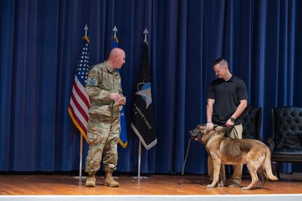 A dog and his handler stand on stage with a military member.