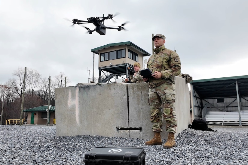 A soldier flies a drone over gravel and concrete under a cloudy sky.