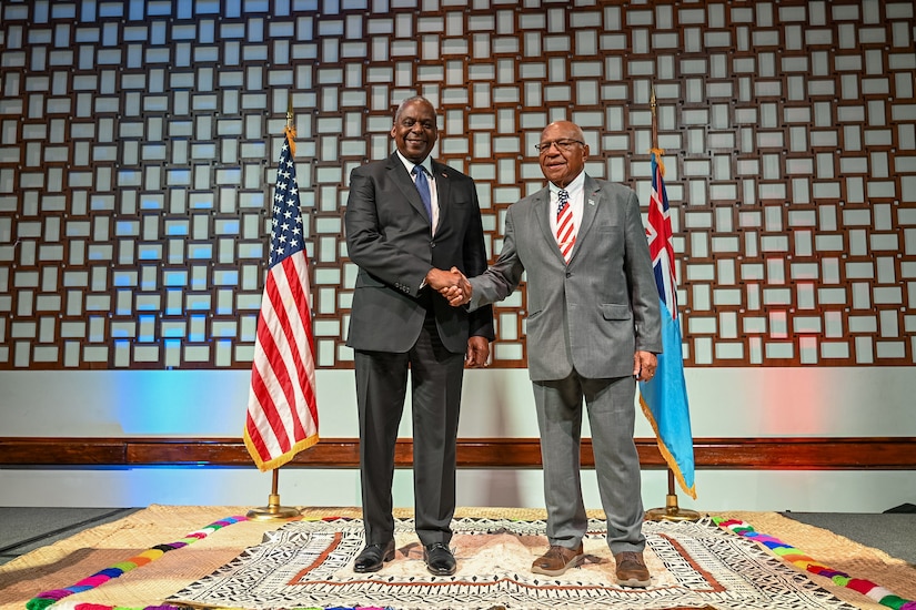 Two men wearing business attire shake hands on a stage.