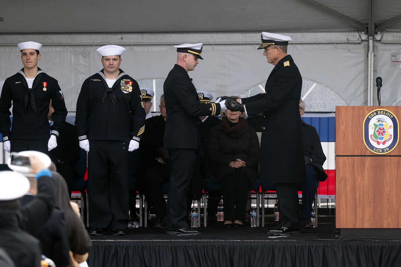 A sailor presents a long glass to another sailor while on a stage with other sailors in front of an audience of sailors.