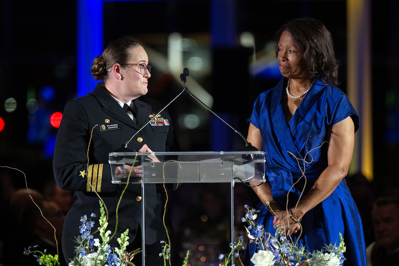 Two high-ranking female service members, one in uniform and one in a blue dress, stand behind a podium and microphone.