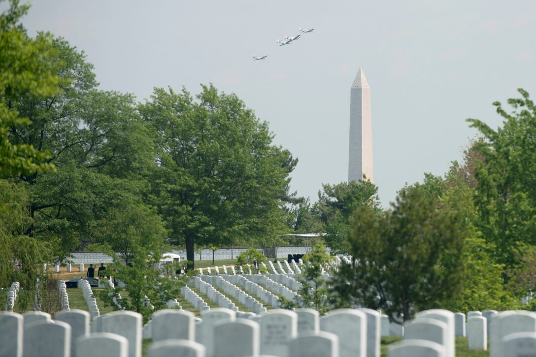 Four aircraft fly in the distance near a tall obelisk. Grave markers in a cemetery are in the foreground.