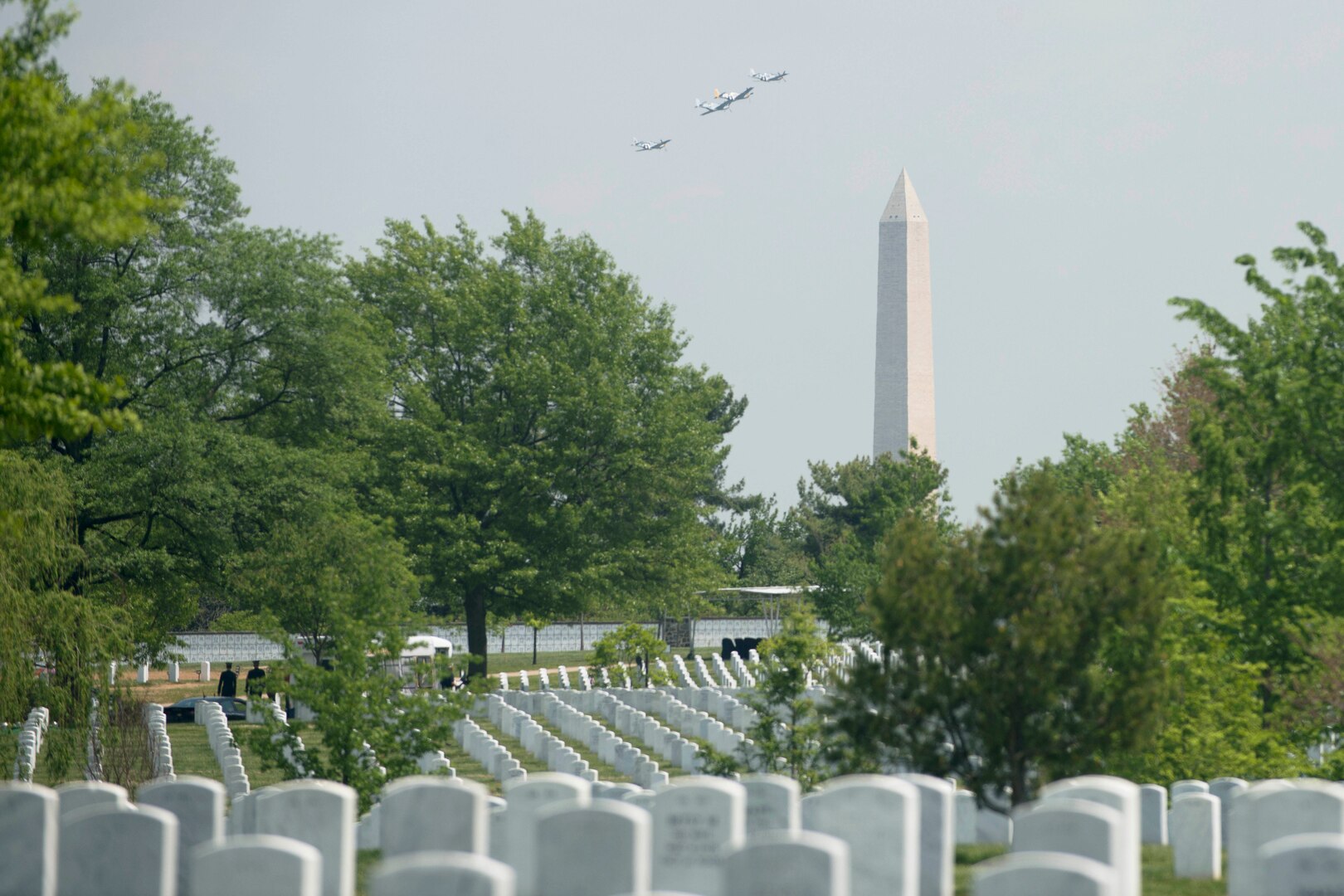 Four aircraft fly in the distance near a tall obelisk. Grave markers in a cemetery are in the foreground.