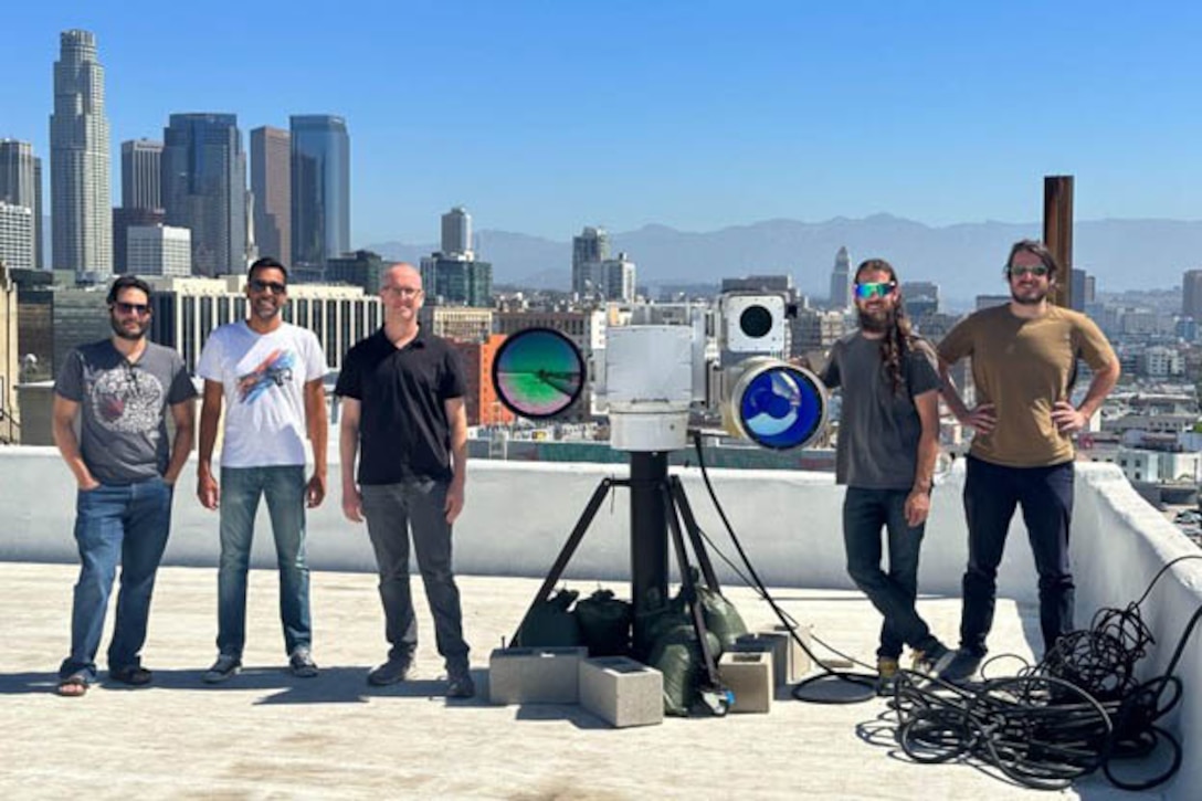 Five men stand around a large camera display on top of a building with a city view in the background.