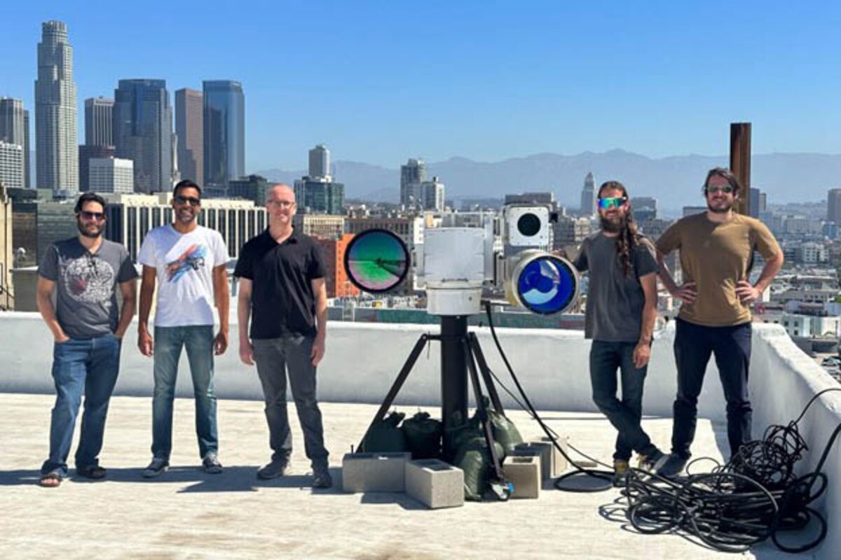 Five men stand around a large camera display on top of a building with a city view in the background.
