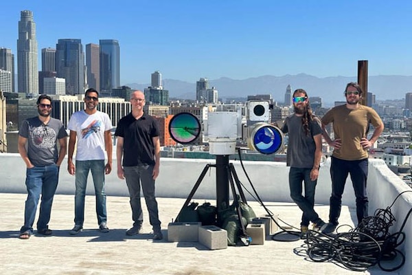 Five men stand around a large camera display on top of a building with a city view in the background.