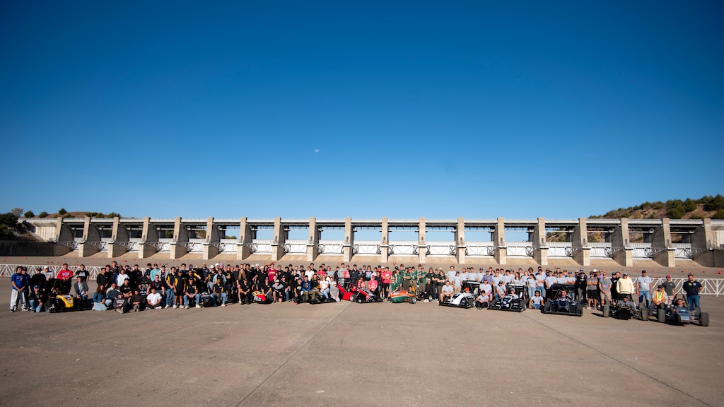 Dozens of people stand behind eight formula one race cars with grey concrete in the foreground and blue sky in the background.