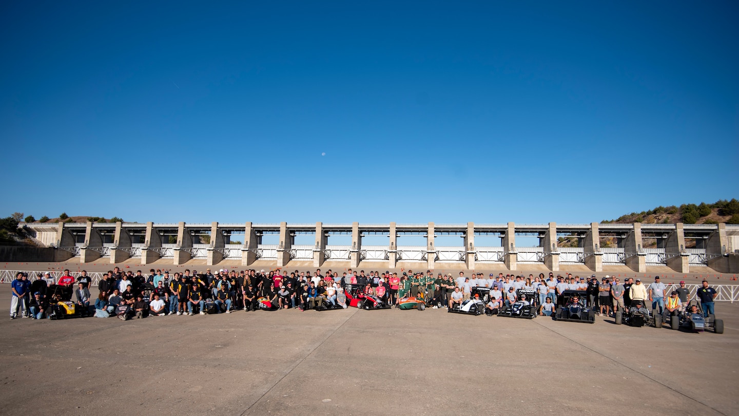 Dozens of people stand behind eight formula one race cars with grey concrete in the foreground and blue sky in the background.