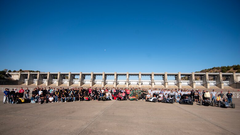 Dozens of people stand behind eight formula one race cars with grey concrete in the foreground and blue sky in the background.