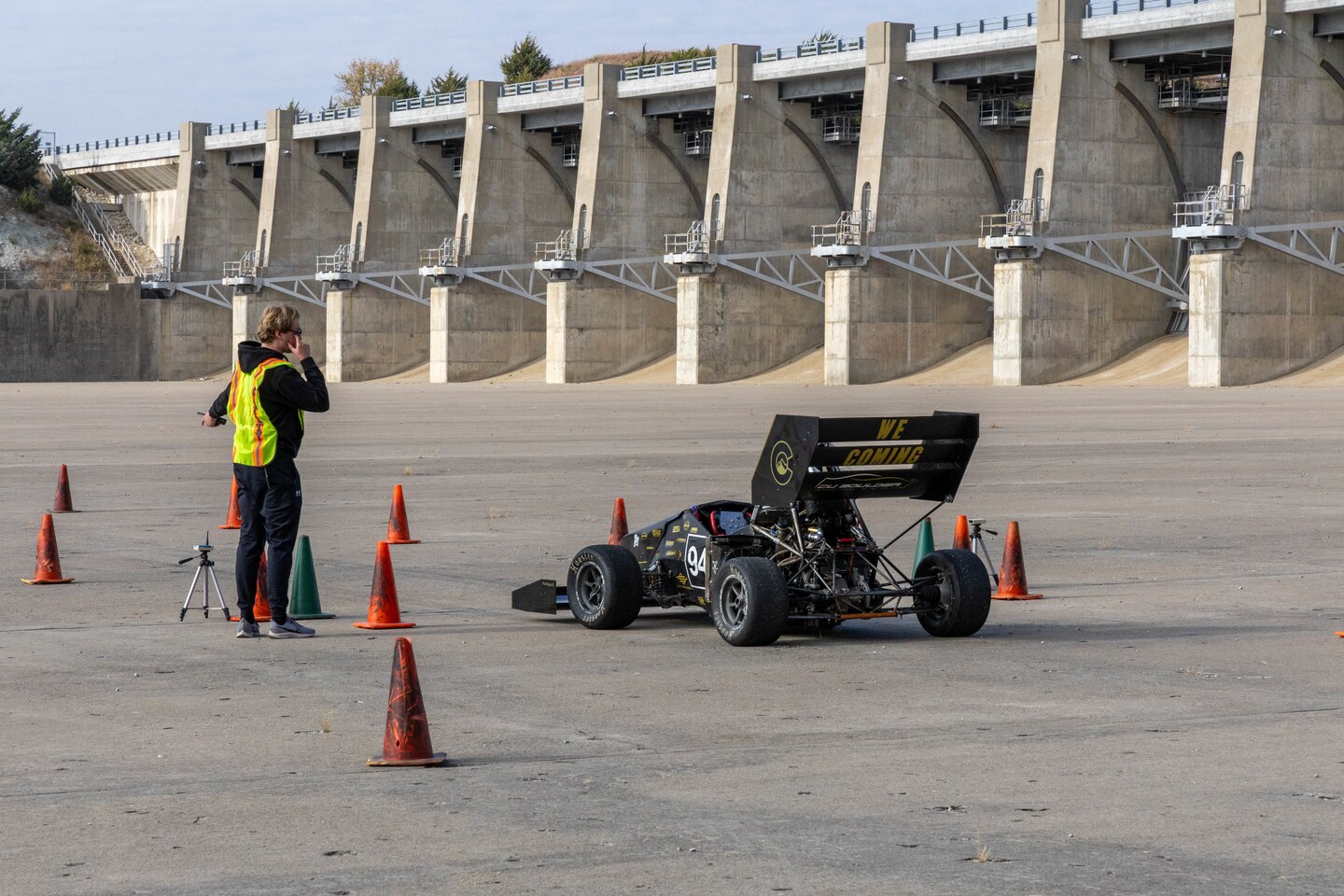 A black formula one race car sits on a grey slab of concrete with orange traffic cones around it and a man in all black with a yellow safety vest to the left.