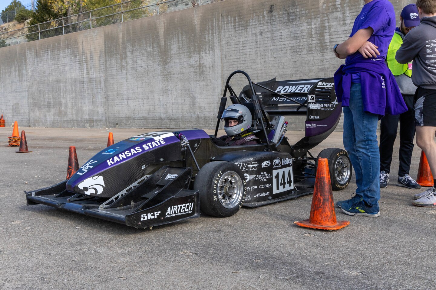 A purple and black formula one race car sits with orange traffic cones around it and a man in a purple shirt and blue pants next to it.