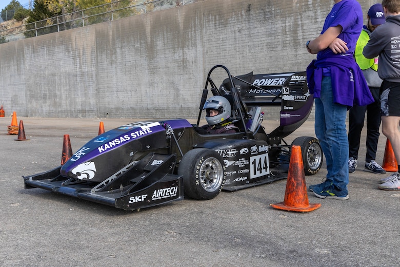 A purple and black formula one race car sits with orange traffic cones around it and a man in a purple shirt and blue pants next to it.