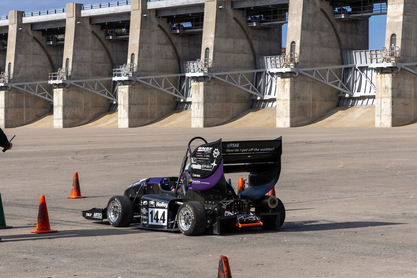 A purple and black formula one race car sits on a grey slab of concrete with orange traffic cones around it.
