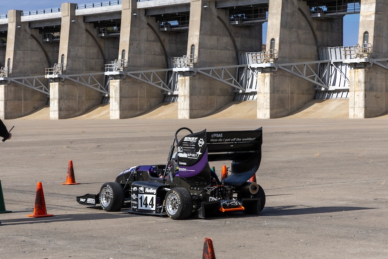 A purple and black formula one race car sits on a grey slab of concrete with orange traffic cones around it.