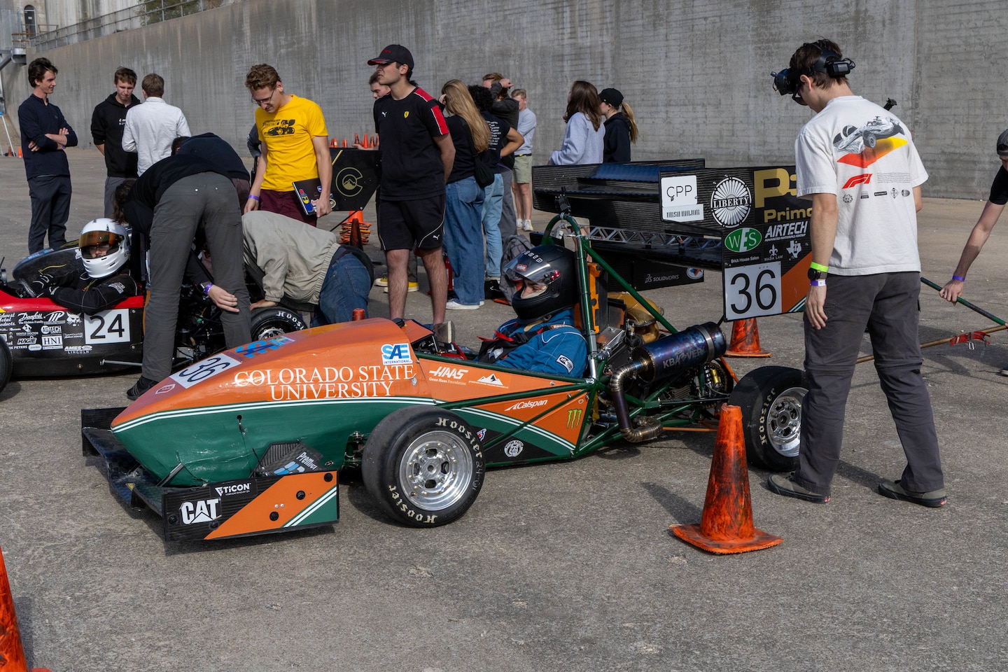 An orange and green formula one race car sits next to an orange traffic cone with a man in a white shirt and grey pants to the right.