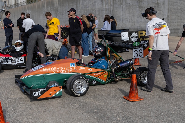 An orange and green formula one race car sits next to an orange traffic cone with a man in a white shirt and grey pants to the right.