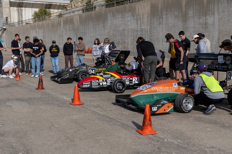 Three formula one race cars are lined up with orange traffic cones in front of them and several people standing behind them.