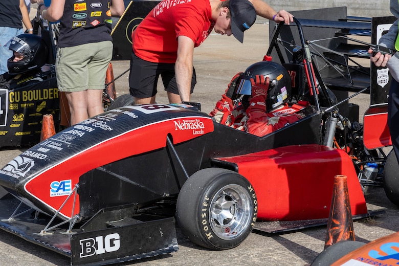 A black and red formula one race car with a driver inside and a man in black shorts with a red shirt bends over the car.