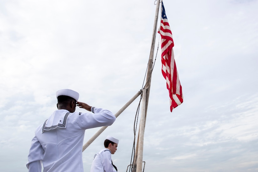 A sailor salutes a U.S. flag outdoors as another handles lines on the flagpole.