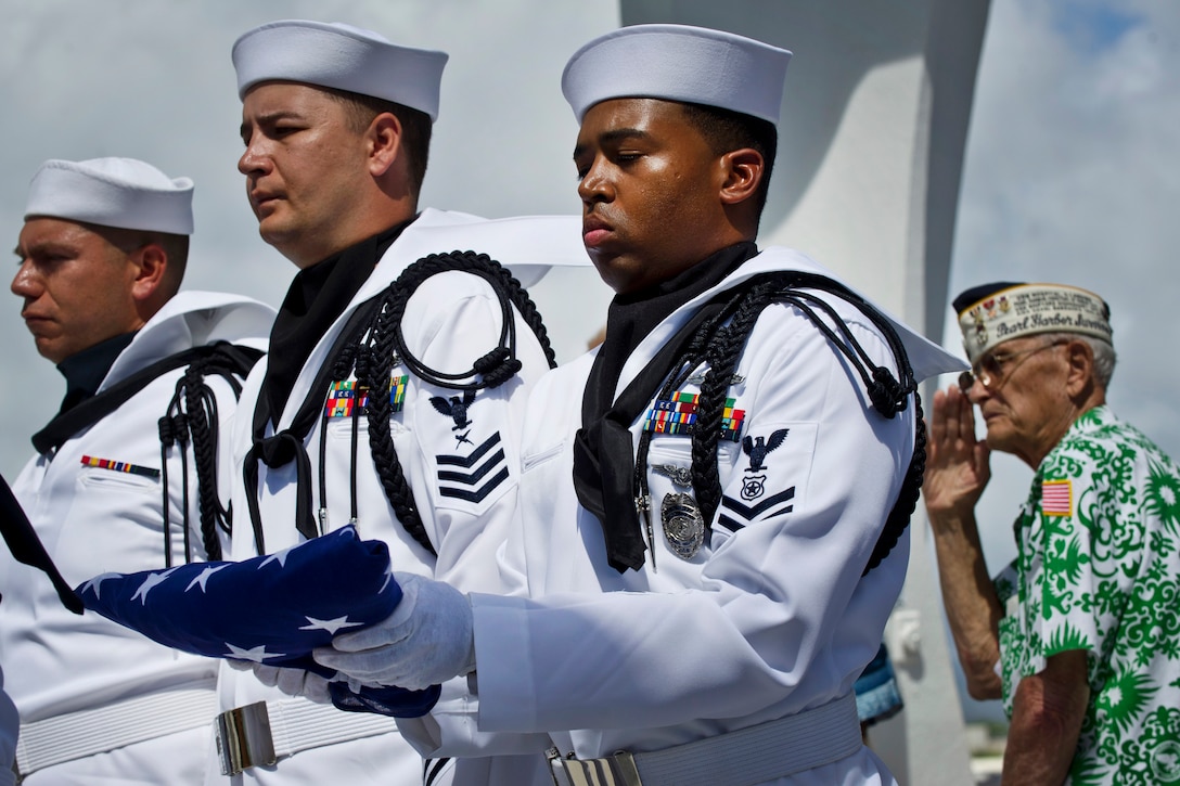 A sailor holds a folded U.S. flag as two others stand nearby while a veteran salutes in the background.