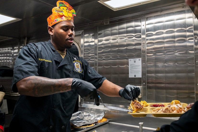 A sailor wearing black gloves and a Thanksgiving headpiece serves food on a tray while in a stainless steel kitchen.