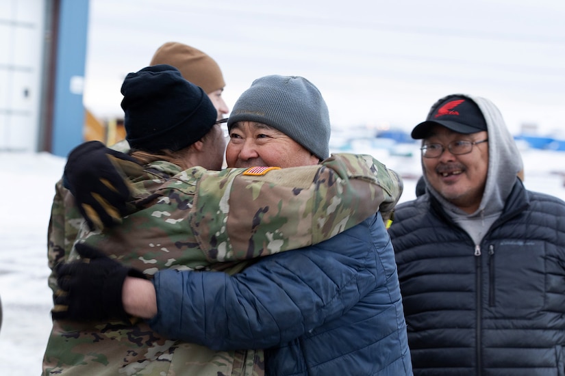 A service member hugs a civilian while outside wearing hats and coats.