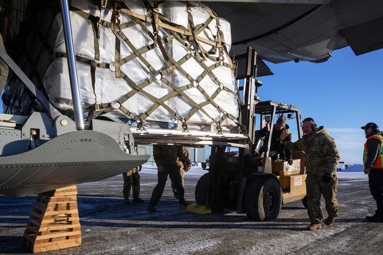 A group of guardsmen use a tractor to remove a large pallet of supplies from the cargo of an aircraft.