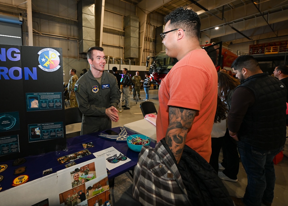 U.S. Air Force Staff Sgt. Aaron Schmidt, 316th Training Squadron airborne cryptologic language analyst instructor supervisor, discusses the 316th TRS mission with a booth visitor during the 42nd annual Santa’s Market and 2nd annual Base Open House at the Louis F. Garland Department of Defense Fire Academy, Goodfellow Air Force Base, Texas, Nov. 23, 2024. The 316th TRS’s mission is to develop leaders and deliver relevant and realistic joint intelligence, surveillance and reconnaissance training for the Department of Defense. (U.S. Air Force photo by Airman 1st Class Evelyn D'Errico)