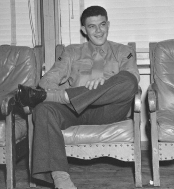 A smiling man sits in a chair with one leg crossed over the other.