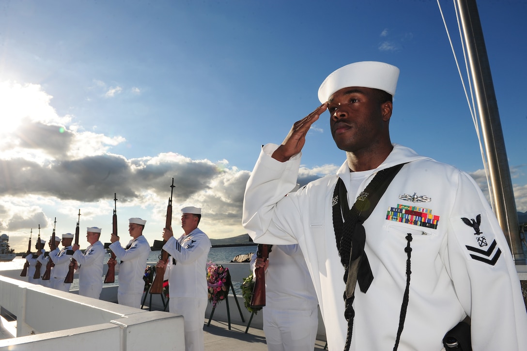 A sailor stands and salutes on an open structure over water, as fellow sailors stand holding weapons in a row nearby.