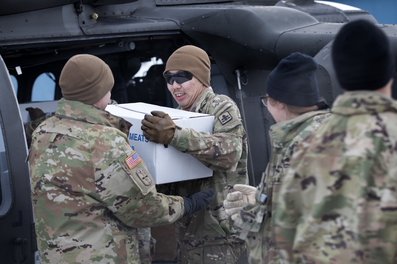 A group of guardsmen offload boxes of meat from a helicopter while wearing hats and gloves.