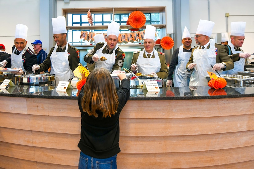 A child holds up a plate for food being served by several military officers behind a counter with Thanksgiving decorations.