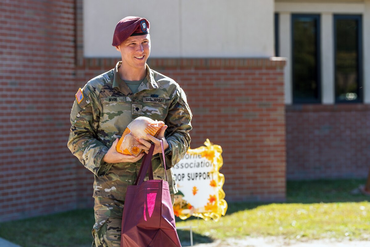 A soldier walks while holding a turkey with a building in the background.