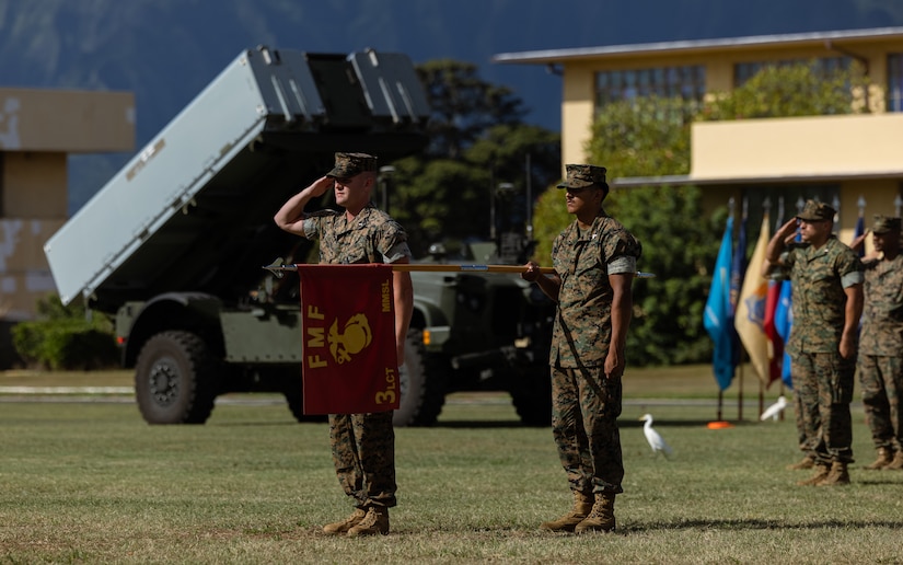 Marines stand in formation on a field near a military vehicle with a weapon system on it.