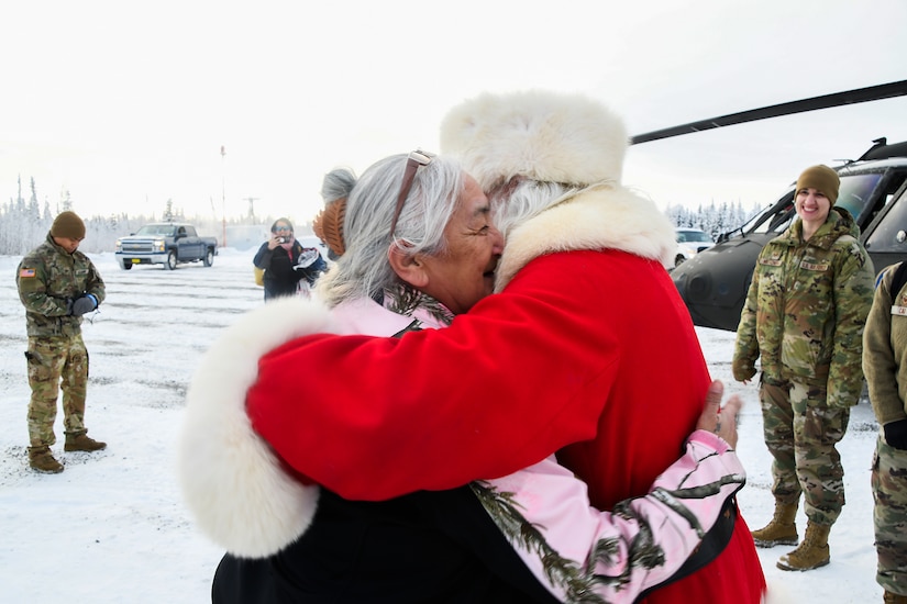 A man dressed in a red robe and white fuzzy hat hugs a woman while outside on snow-covered field.