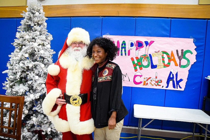 Santa Claus smiles for a photo with another person in a gym-type room next to a Christmas tree.