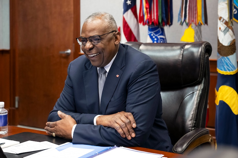 The secretary of defense smiles while sitting at a desk with flags visible in the background.