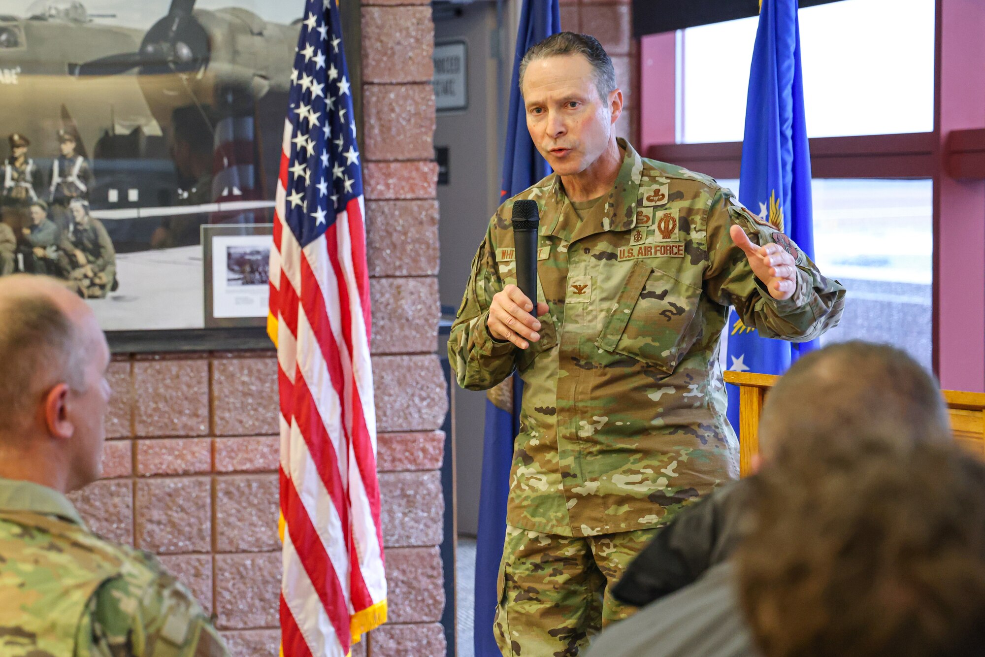 Uniformed Air Force officer stands in front of crowd, holding microphone.