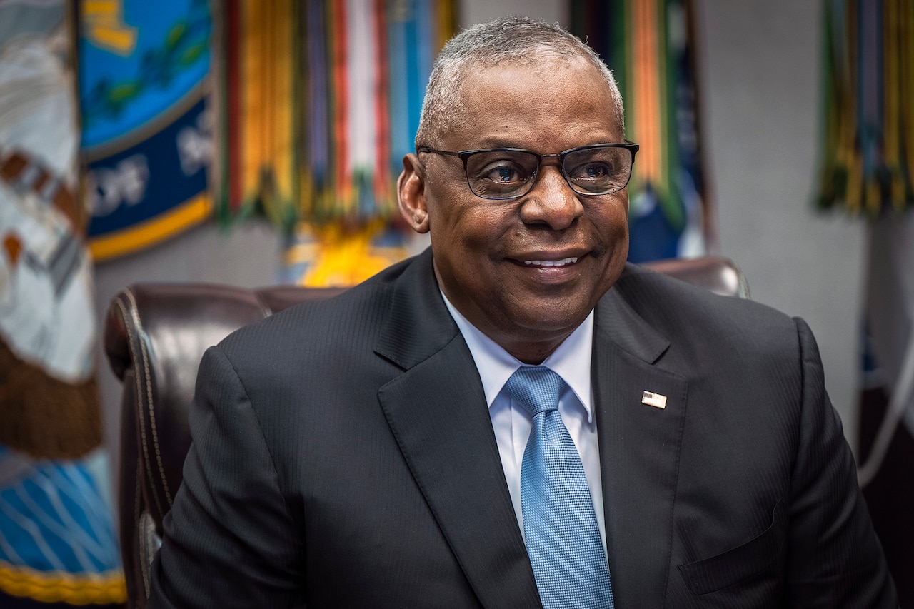 The secretary of defense smiles while sitting at desk in front of flags.