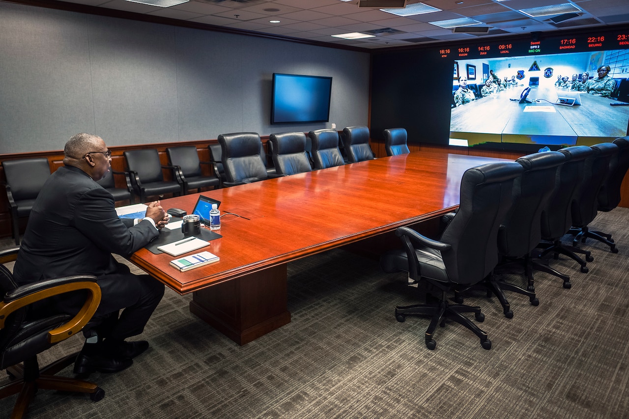 The secretary of defense looks at a screen while sitting at the end of a conference table.