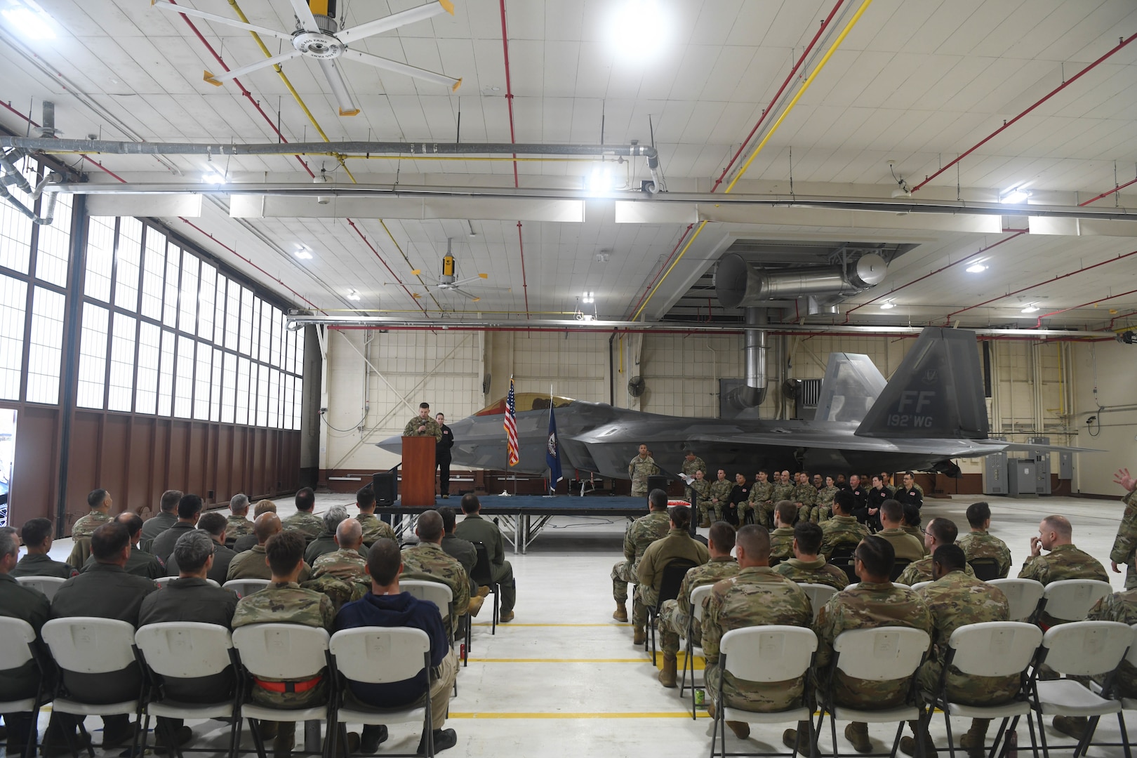 View of the audience sitting in chairs with a stage and F-22 Raptor in the background.