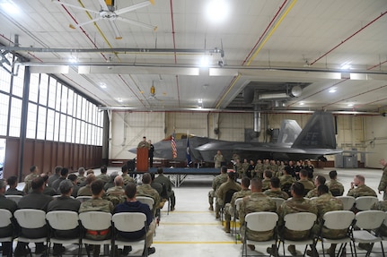 View of the audience sitting in chairs with a stage and F-22 Raptor in the background.